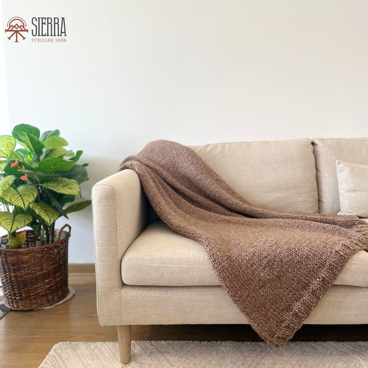 A brown knitted blanket draped over the back of a beige sofa, with a plant basket on the side, set in a well-lit room.