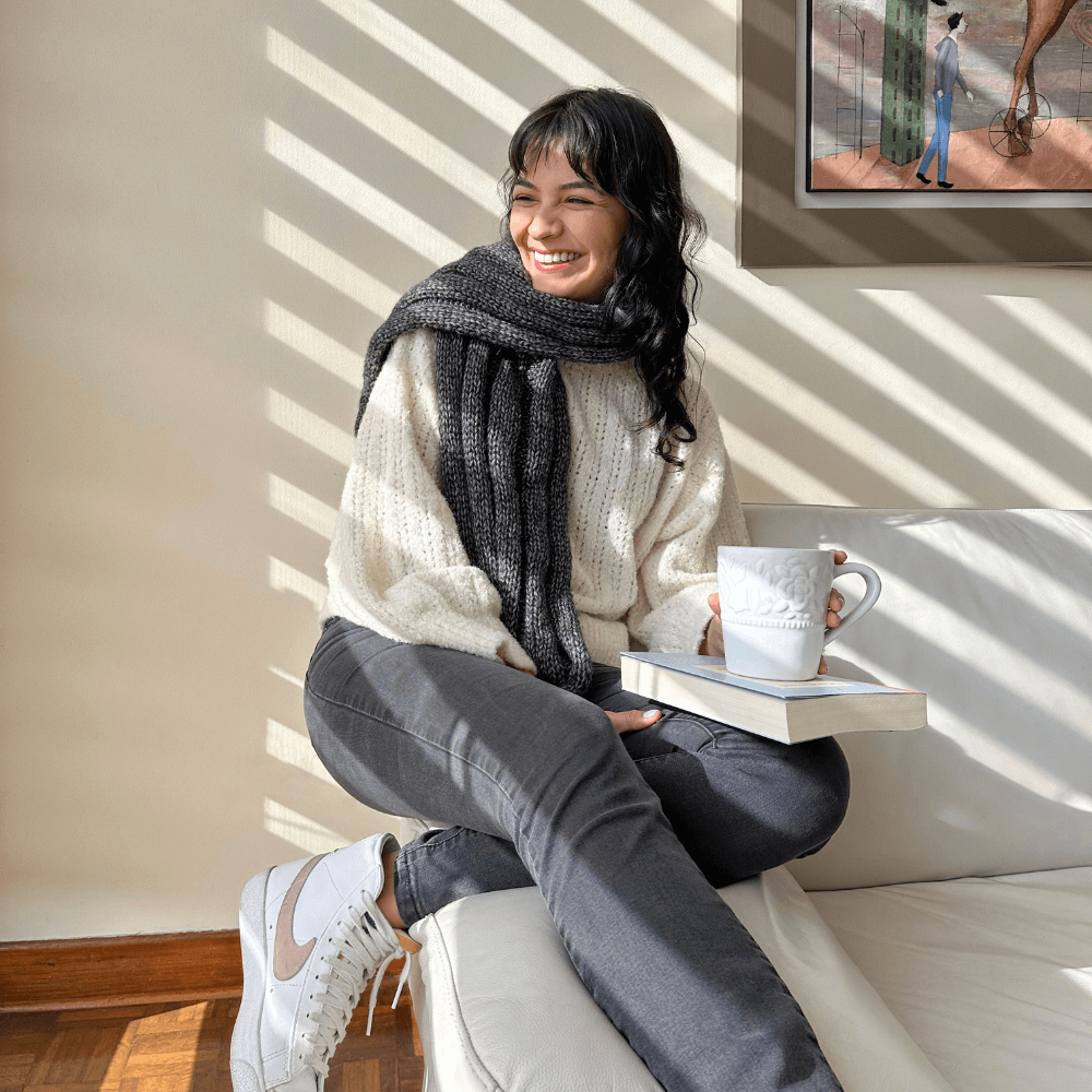 A person sitting on a sofa wearing a beige sweater and gray pants, knitting a gray scarf using knitting needles. A cup of tea and a book are also visible on the sofa.