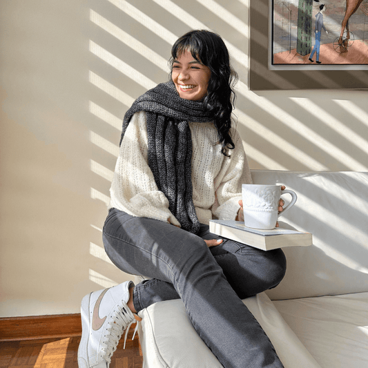 A person sitting on a sofa wearing a beige sweater and gray pants, knitting a gray scarf using knitting needles. A cup of tea and a book are also visible on the sofa.
