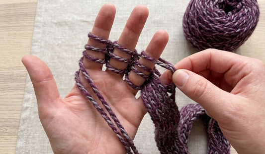 How to finger knit demonstration with yarn woven over four fingers and a finished knitted cord growing from the hand next to a ball of purple marled yarn