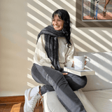 A person sitting on a sofa wearing a beige sweater and gray pants, knitting a gray scarf using knitting needles. A cup of tea and a book are also visible on the sofa.