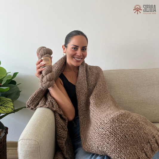 Woman sitting on a couch wearing a brown knitted blanket, holding yarn, with a plant and light-colored wall in the background.