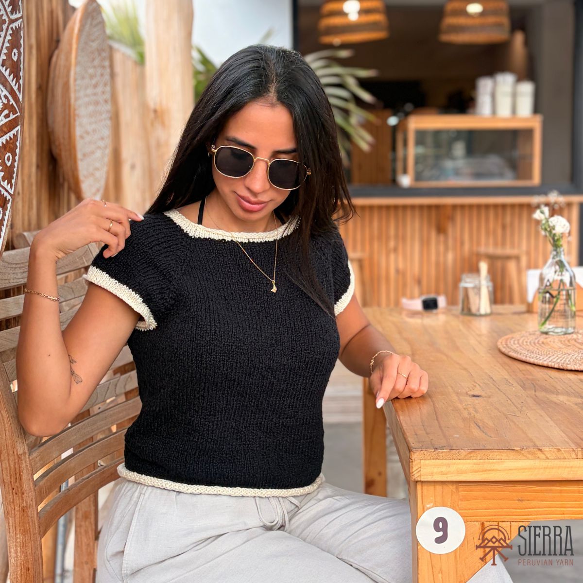 A woman wearing a black short-sleeve knit top with cream-colored trim, paired with light gray pants, sitting at a wooden table in a rustic cafe setting.