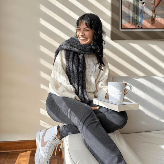 A person sitting on a sofa wearing a beige sweater and gray pants, knitting a gray scarf using knitting needles. A cup of tea and a book are also visible on the sofa.