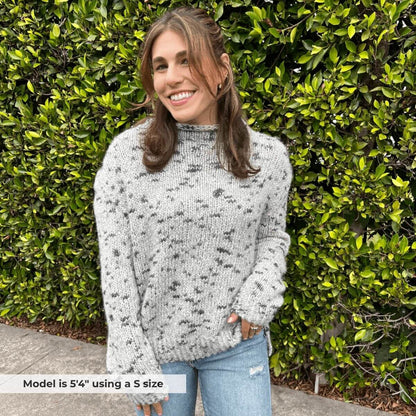 Woman wearing a gray patterned sweater and blue jeans standing in front of green bushes.