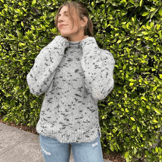 A person wearing a gray speckled pullover with a round neckline standing in front of a hedge.